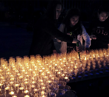 Nighttime photo with a tabletop covered in lit tealight candles and a few students lighting candles.